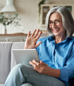 Happy 60s older mature middle aged adult woman waving hand holding digital tablet computer video conference calling by social distance virtual family online chat meeting sitting on couch at home.